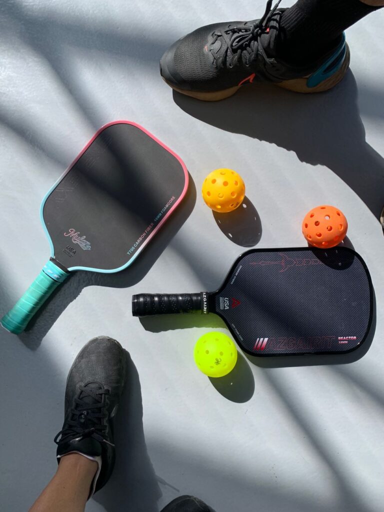 Two paddles and balls on a sunlit outdoor pickleball court with players ready to start