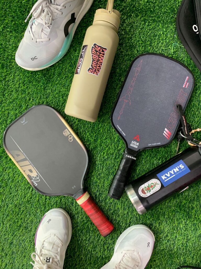 Pickleball paddles and gear on turf, one paddle showing the USA Pickleball approved stamp tied to the pickleball spin test ball standard