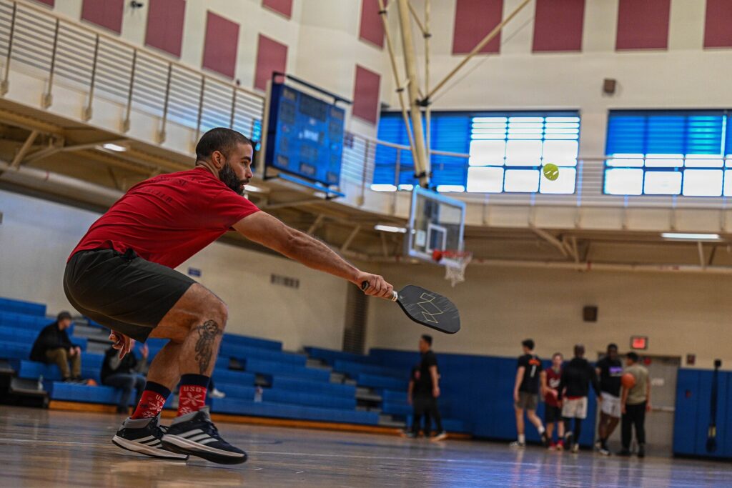 Player reaching low for a pickleball in a gym, used to illustrate a guide to Ben Johns age and career timeline