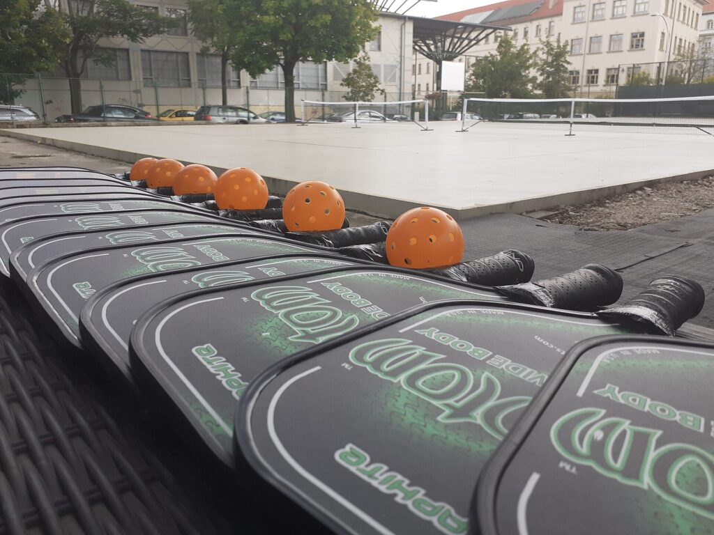A row of identical widebody pickleball paddles and orange balls beside an outdoor court, the kind of older model that ends up among discontinued pickleball paddles