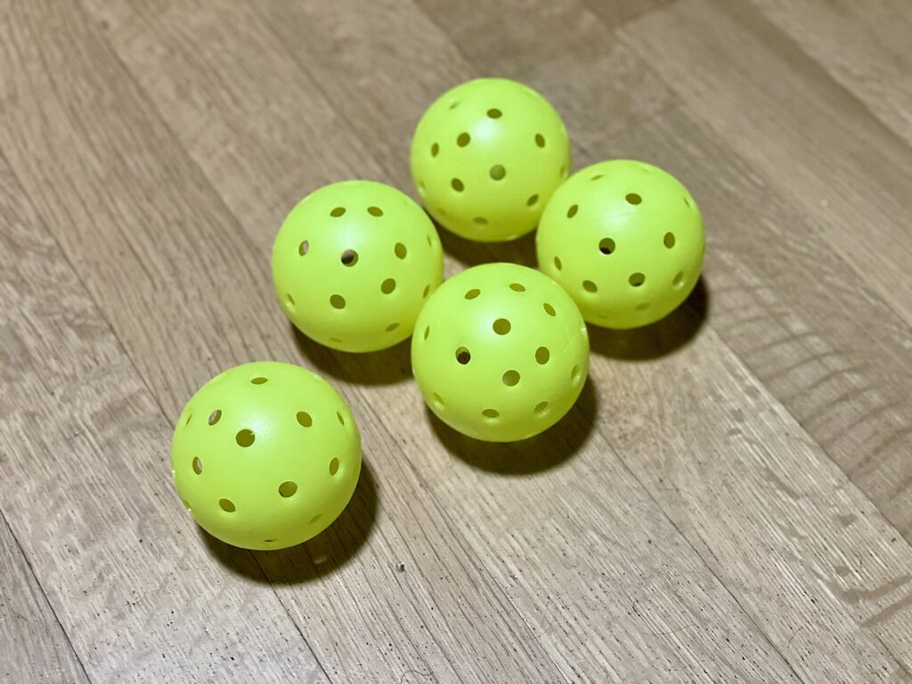 Five unbranded yellow pickleballs on a wooden floor, the kind of gear to check along with fake pickleball paddles