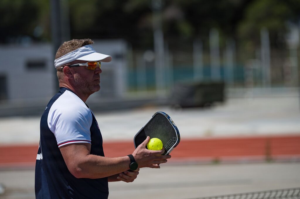 Pickleball player in a visor holding a paddle and yellow ball between points on an outdoor court
