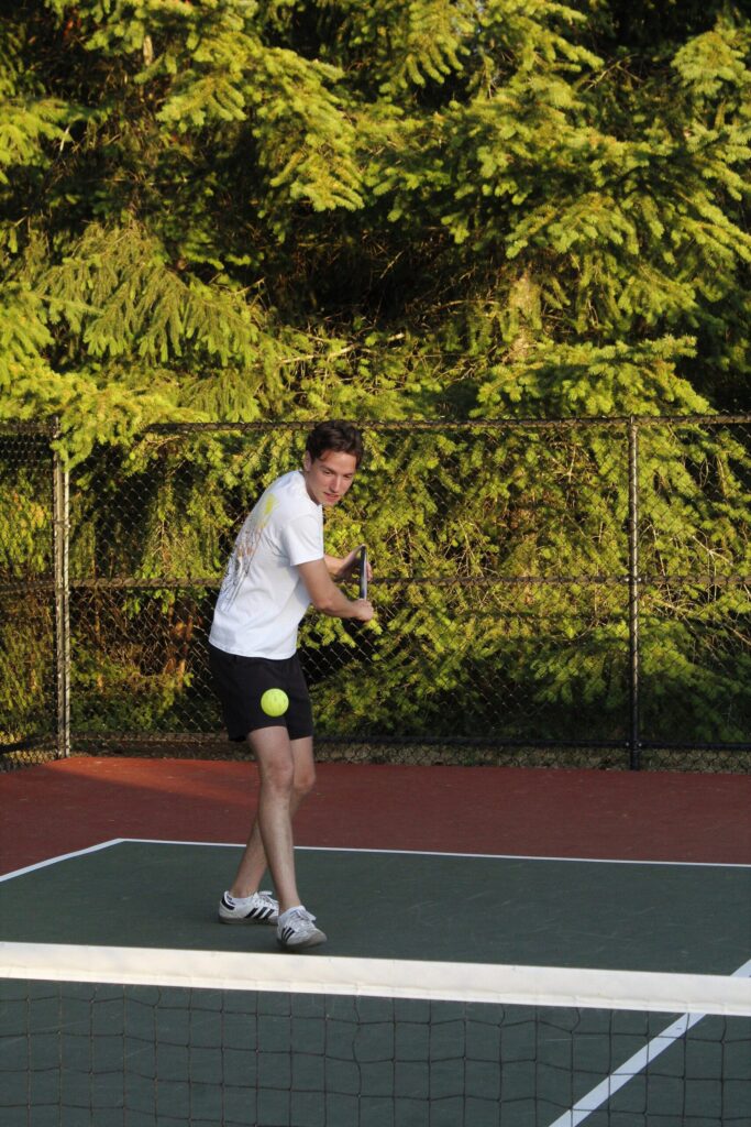 Player turning to hit a backhand on an outdoor court lined with evergreens, the shot where some players switch hands in pickleball